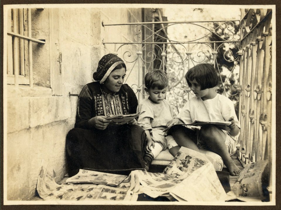 Arab woman in traditional dress, seated with boy and girl (possibly members of the American Colony), looking at comics LCCN2007675266