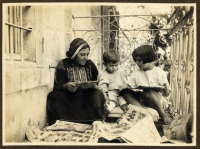 Arab woman in traditional dress, seated with boy and girl (possibly members of the American Colony), looking at comics LCCN2007675266