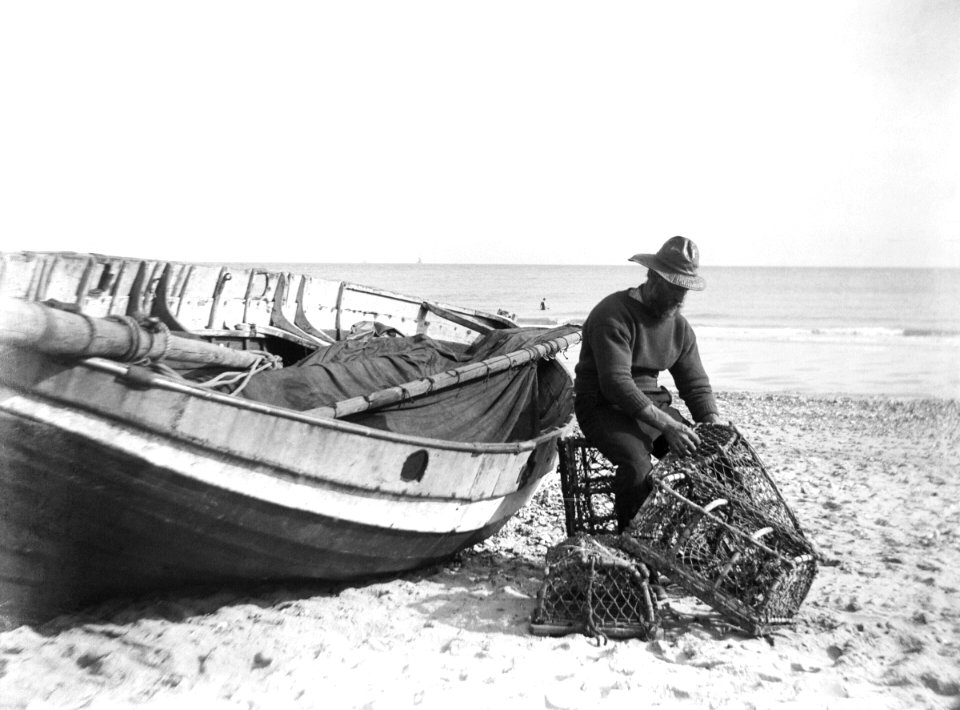A fisherman in sou'wester mending lobster-crab creels on the beach alongside a beached Sheringham crab boat. RMG P27501
