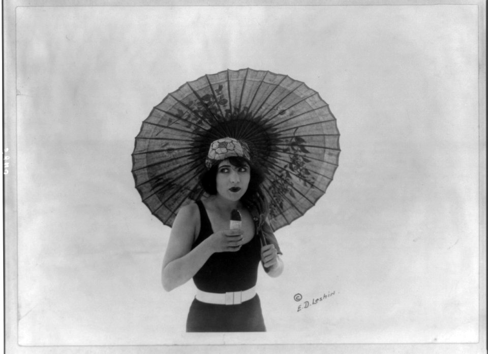 Betty Compson, half length portrait, standing, with umbrella, in bathing suit, facing slightly right, eating Eskimo Pie. Movie star LCCN2005691986