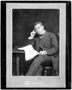 Booker T. Washington, three-quarter length portrait, seated, facing slightly left, holding newspaper LCCN98500584