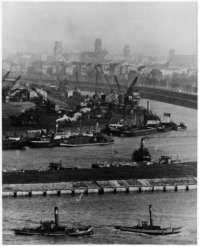 Duisburg, Germany. The camera looks across the Rhine river to the hazy towers of Duisburg. Busy tugs, loading cranes... - NARA - 541698