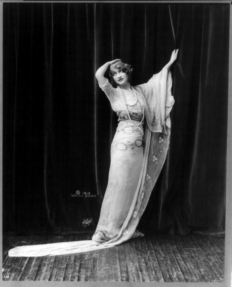 Gaby Deslys, 1884-1920, full length portrait, standing, facing left, rt. hand on top of head, lt. hand grasping curtain LCCN2005693306