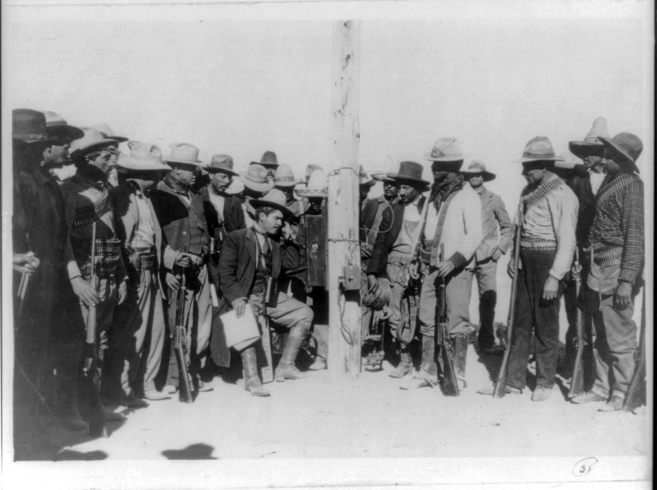 Gen. Campa, full, seated, talking on telephone attached to pole; surrounded by his troops LCCN2007677276