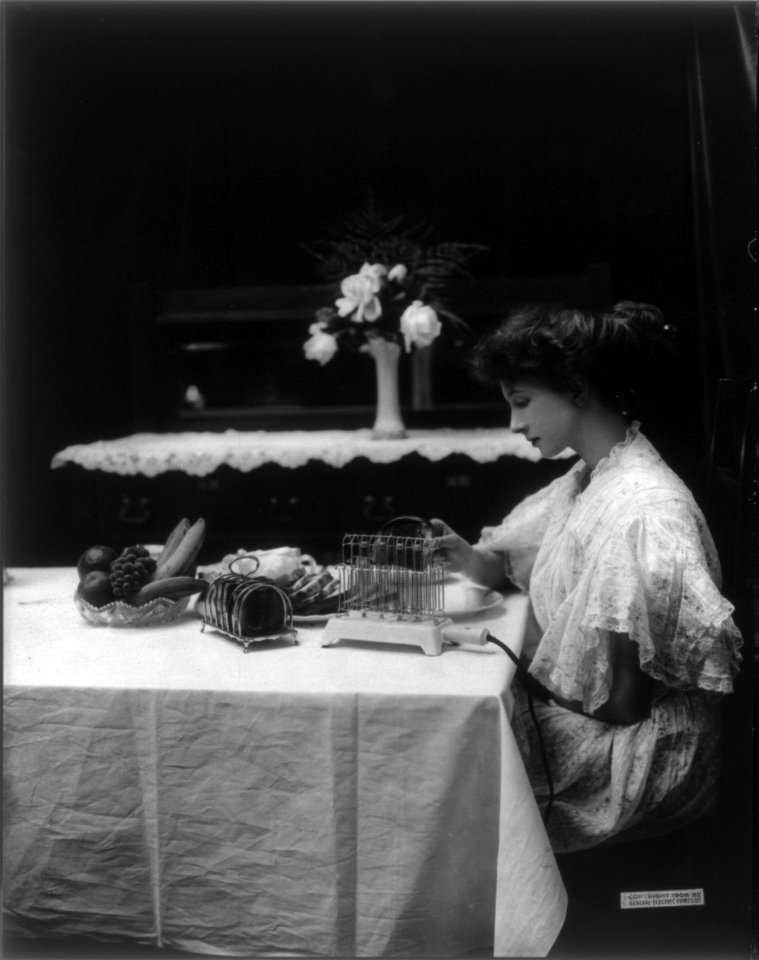 Gene and her friend using General Electric Heating and Cooking Devices- young woman seated at table, removing toast from toaster LCCN2006677998