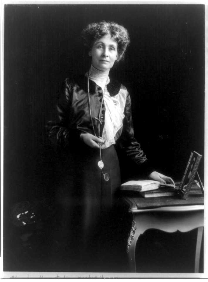Emmeline (Goulden) Pankhurst, three-quarter length portrait, standing by small table, facing right, with left hand on book LCCN96510315