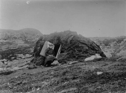 En kvinne sitter i døråpningen til en torvgamme, Skogerøya, Sør-Varanger, Finnmark, 1890-årene - Norsk folkemuseum - NF.05063-023