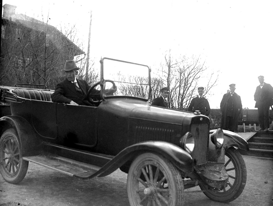 En man sitter i en bil invid järnväg. Fyra uniformsklädda män i bakgrunden - Nordiska Museet - NMA.0054299