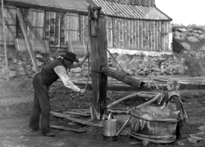 En man som pumpar vatten och en liten flicka vid en gammal pump. Ca 1880-1890. Eriksbergsområdet
