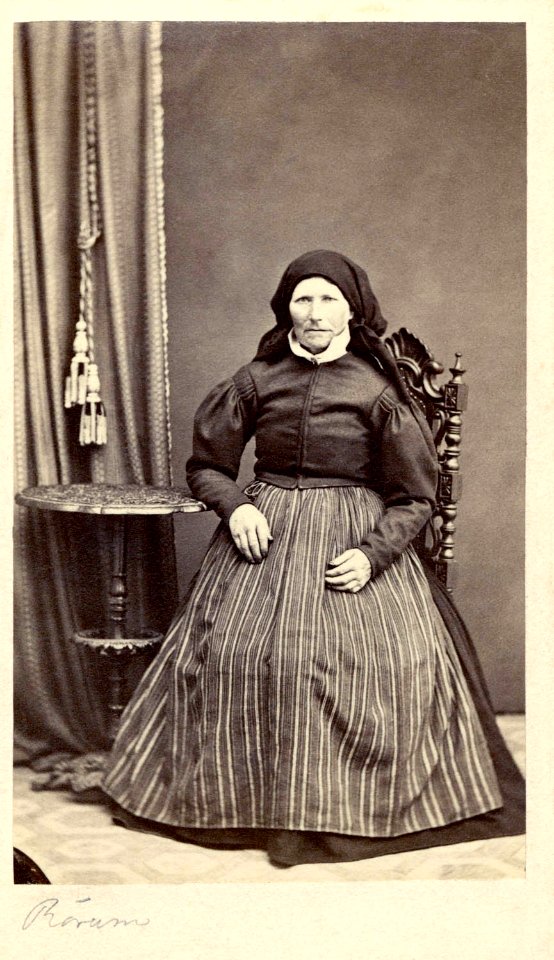 En äldre kvinna i schalett sitter på en stol och vilar armen på ett litet pelarbord. Rörum - Nordiska Museet - NMA.0039727