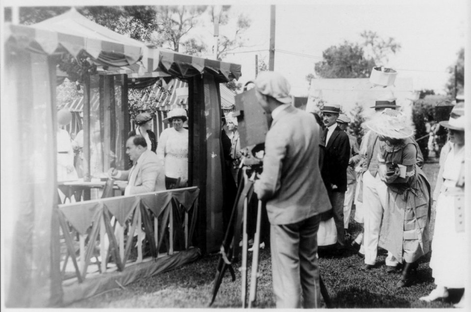 Enrico Caruso, 1873-1921, drawing caricature sketches in booth at charity fair in Southampton, L.I., left profile, showing exterior of booth, onlookers and photographer at work LCCN2005680290
