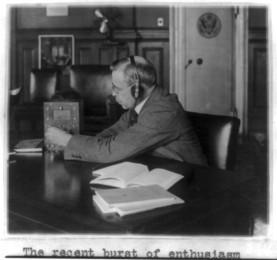 Government official seated at his desk wearing head phones and tuning small radio LCCN2002697154