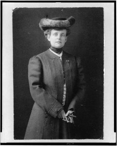 Frances Benjamin Johnston, half-length portrait, standing, facing front, wearing hat and holding gloves LCCN95506164