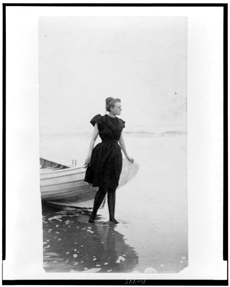 Frances Benjamin Johnston, full-length portrait, standing at edge of ocean, in bathing suit, with left hand on boat, facing right LCCN98506467