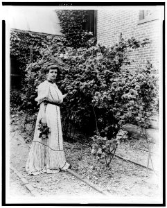 Frances Benjamin Johnston, full-length portrait, standing in her garden, by rose bushes, with some flowers in her hand, facing slightly right LCCN98506473