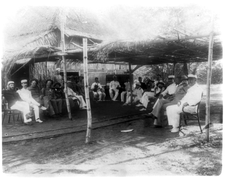 Group of American soldiers posed under thatched-roof outside, piano and man with violin, San Roque (Cavite), Philippines LCCN2002724002