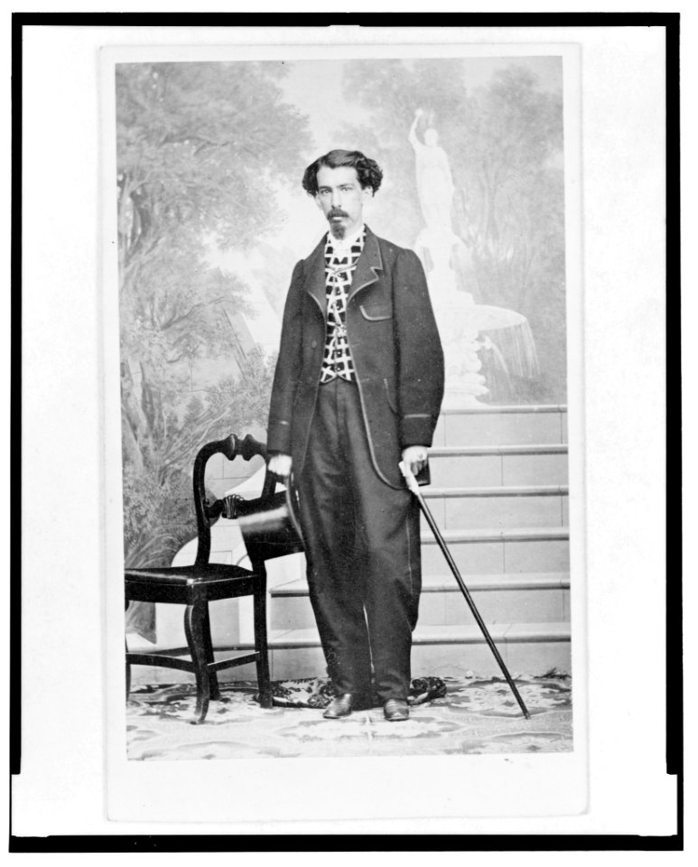 Full-length portrait of a man holding top hat and cane, standing beside chair, facing front, stairs and mural in background LCCN2005692504