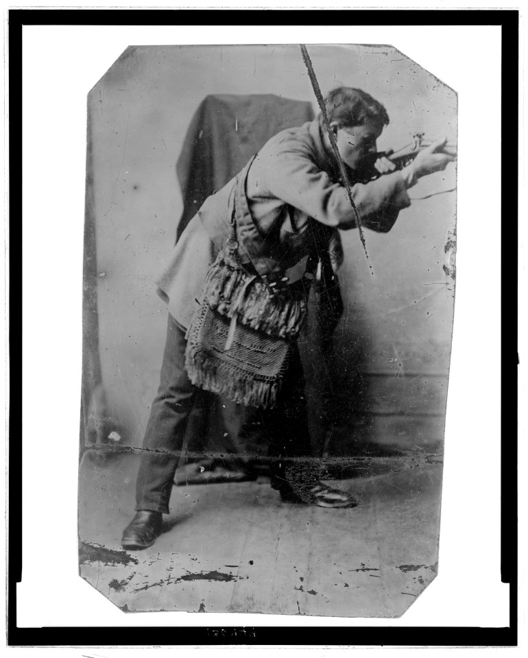 Full-length portrait of a man with a fringed, knitted bag across his chest, posed as if shooting a rifle LCCN2006689570