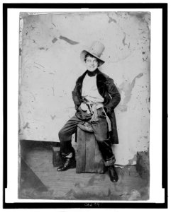 Full-length portrait of a man, dress in frock coat and top hat with sash and pistol at his waist, seated on a crate LCCN2006689590