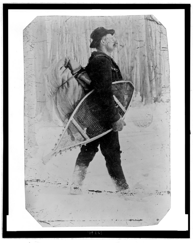Full-length portrait a man carrying a bundle on his back and snowshoes under one arm, appearing to walk through a snowy landscape LCCN2006689563
