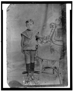 Full-length portrait of a young boy standing next to a wicker chair LCCN2006689594