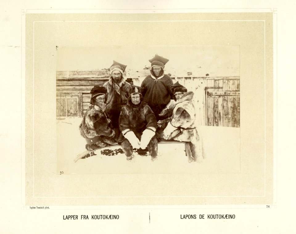 Gruppe med samer fra Kautokeino, sitter på en pulk i snøen kledd i pesk og skaller - Norsk folkemuseum - NF.15006-077
