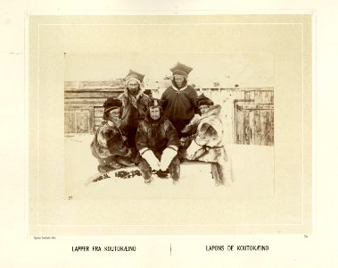 Gruppe med samer fra Kautokeino, sitter på en pulk i snøen kledd i pesk og skaller - Norsk folkemuseum - NF.15006-077
