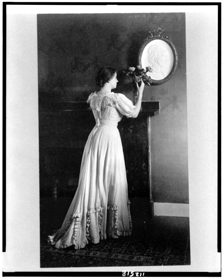 Helen Keller, full-length portrait, facing right, standing at fireplace with flowers on the mantle LCCN94515625