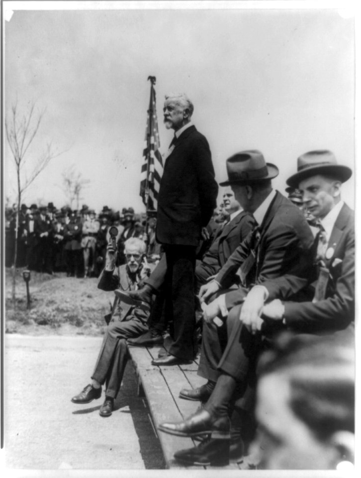 Henry Cabot Lodge standing on platform outdoors making a speech; man seated on platform with hearing aid or recording device LCCN89706583
