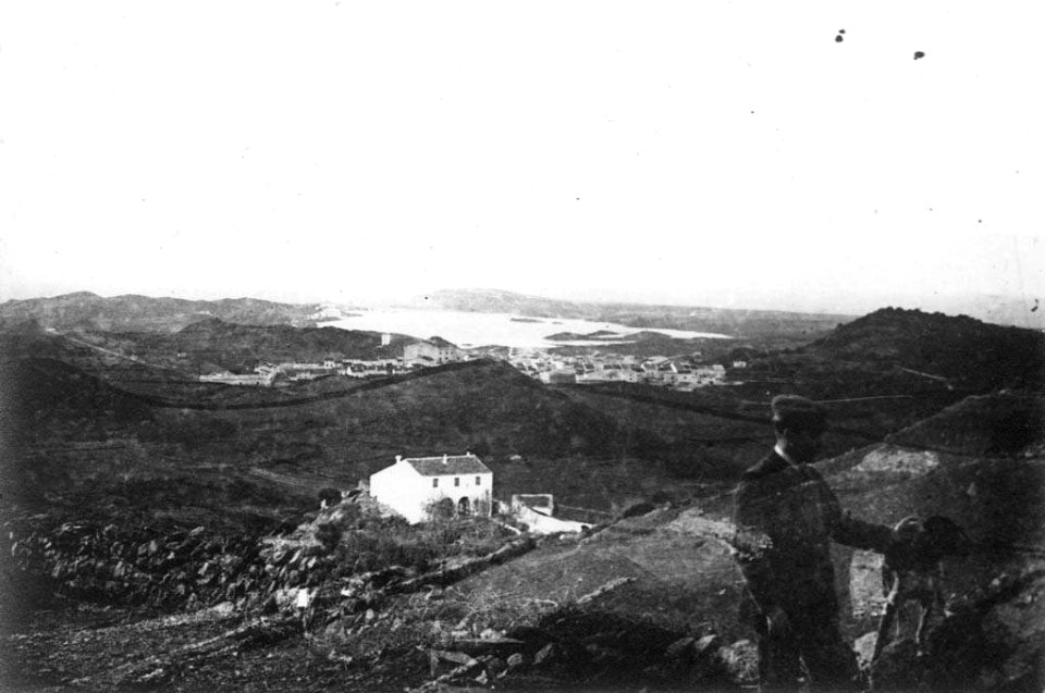 Home amb un gos a la muntanya de Maó i una masia al fons