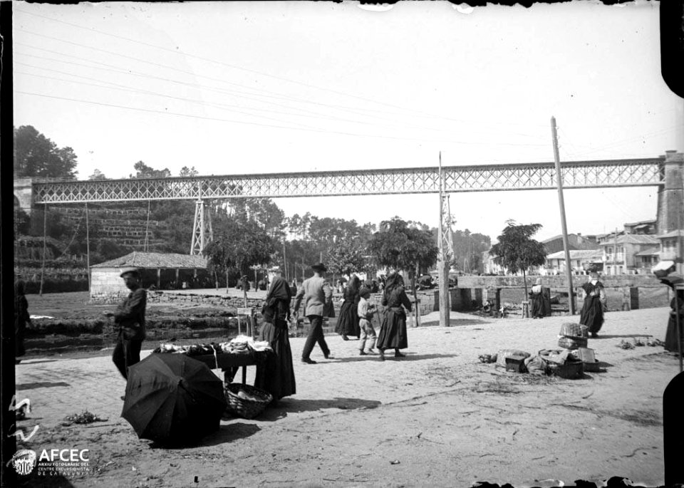 Homes i dones a la riba del riu amb algunes parades i al fons cases i un viaducte