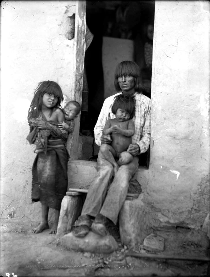 Hopi Indian father sitting in the wood framed doorway of his adobe house with three children, Oraibi, Arizona, ca.1898 (CHS-4579)