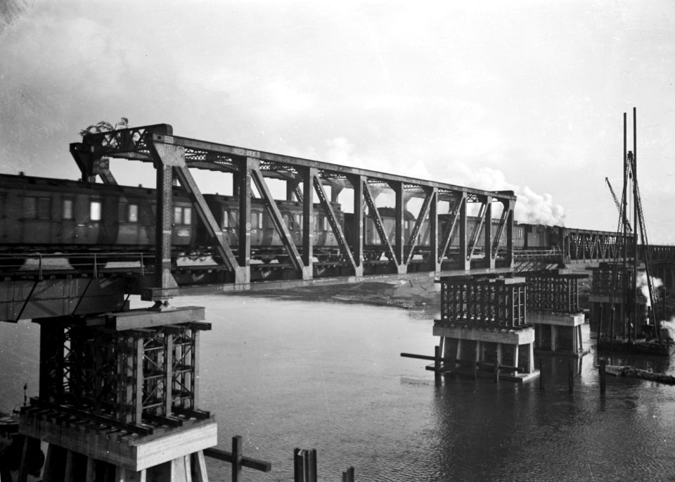 HUA-154934-Gezicht op de herstelde spoorbrug (noodbrug) over de Maas te Buggenum met een stoomtrein