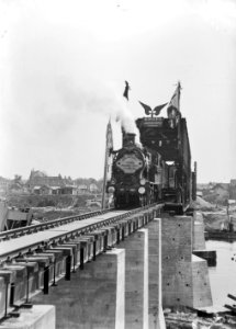 HUA-153627-Gezicht op de nood-spoorbrug over de Maas bij Venlo met een openingstrein getrokken door stoomlocomotief nr. 6011 (serie 6000) van de N.S
