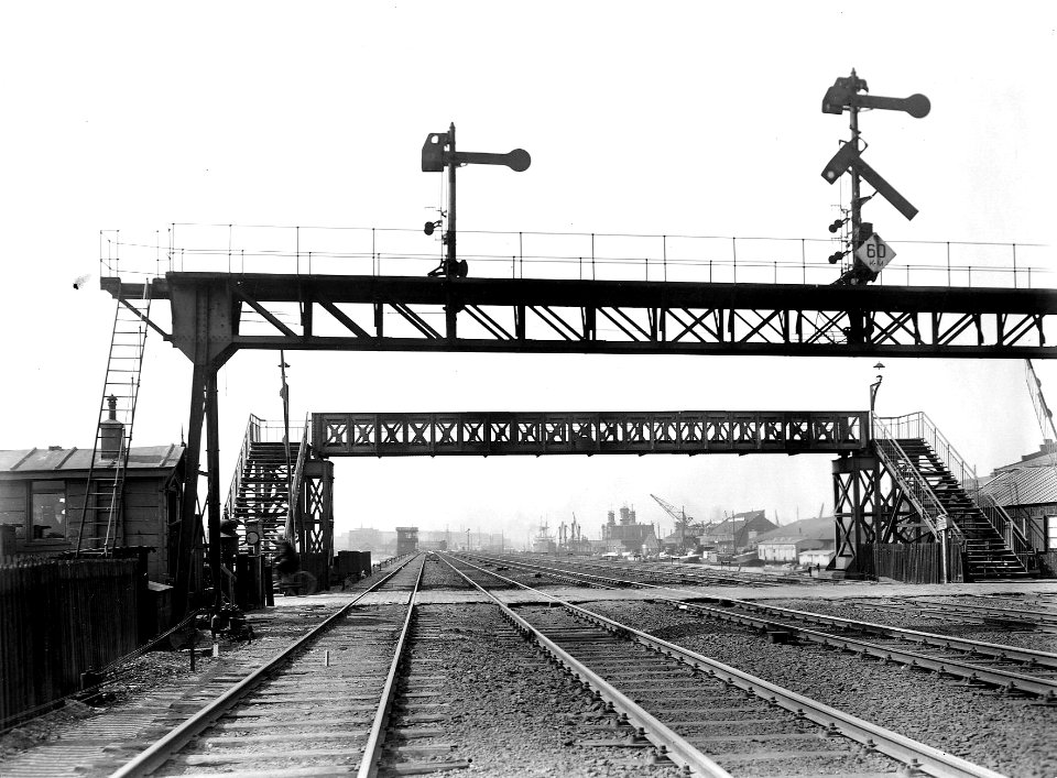 HUA-163644-Gezicht op de spoorwegovergang en de voetgangersbrug richting het Mariniersplein te Amsterdam, ter hoogte van de Handelskade. Op de voorgrond een deel van de seinbrug. N.B. De afbeelding maakt deel ui