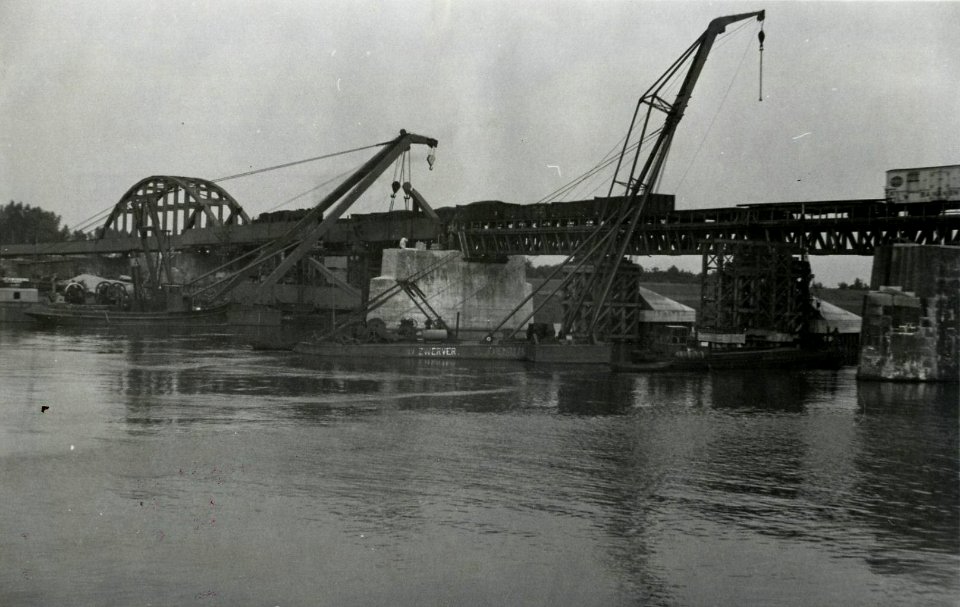 HUA-163527-Gezicht op de werkzaamheden ten behoeve van het herstel van de tijdens de oorlog vernielde spoorbrug over de Maas te Ravenstein, met de noodbrug tijdens het passeren van een goederentrein