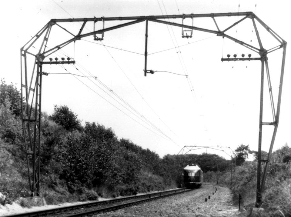 HUA-167122-Gezicht op de van oorlogsschade herstelde (maar nog wel enkelsporige) spoorlijn bij Overveen, met symmetrische bovenleidingsportalen en een electrisch treinstel mat. 1936 van de N.S