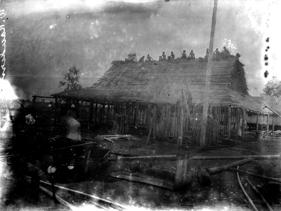Husbygge. En skara infödingar sitter uppe på takryggen. S-te Marie Marovoay. Madagaskar - SMVK - 021870