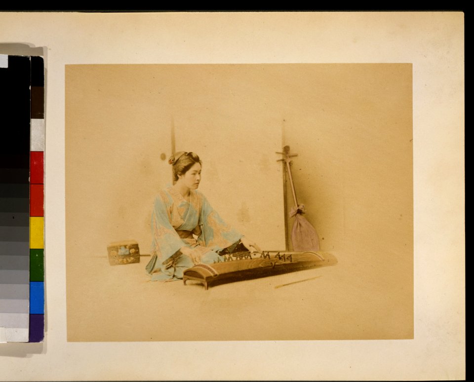Japanese woman, full length, seated, facing right, playing a koto; a shamisen leans against a wall in the background LCCN2009632890