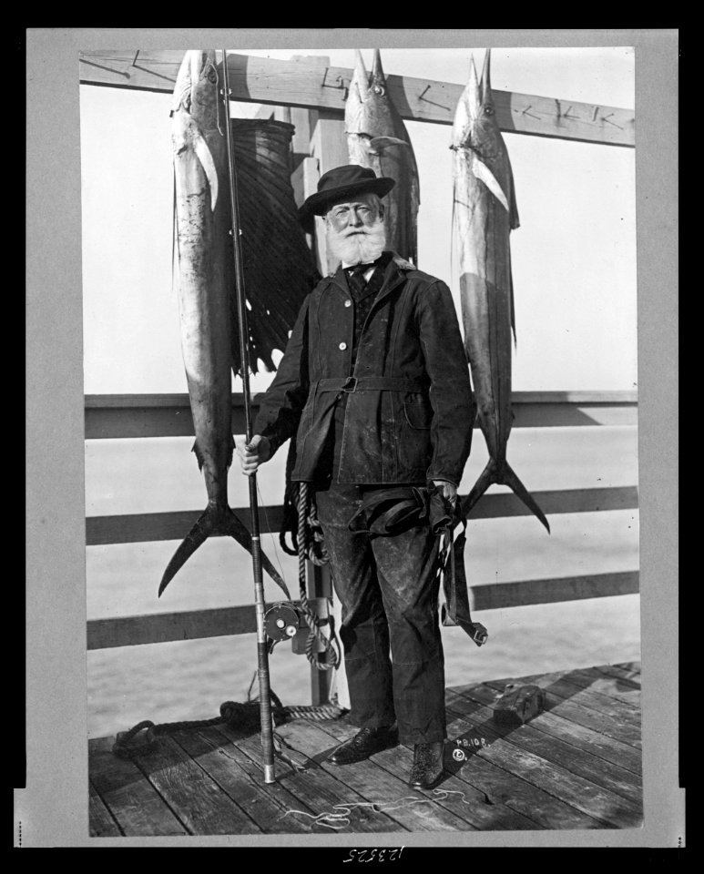 John D. Crimmins, full-length portrait, standing on pier, holding fishing rod and reel, facing front, with three saltwater fish hanging on railing LCCN99471948