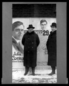 John McCormack, Irish-born American tenor, full-length portrait, standing in front of poster announcing an upcoming performance by him, facing front, with another man LCCN99471543