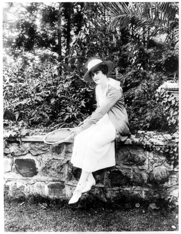 M.E. Kahn, full-length portrait, facing left, seated on stone wall, holding tennis racket LCCN96503932