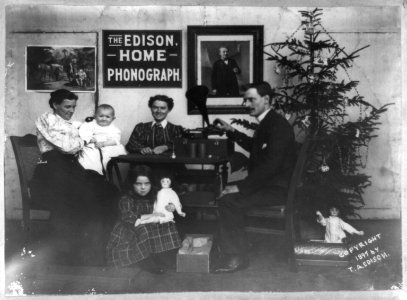 Man, two women and two children listening to phonograph-Girl is holding doll and another doll is under Christmas tree-There is a sign on the wall, The Edison Home Phonograph, and a portrait LCCN97504441