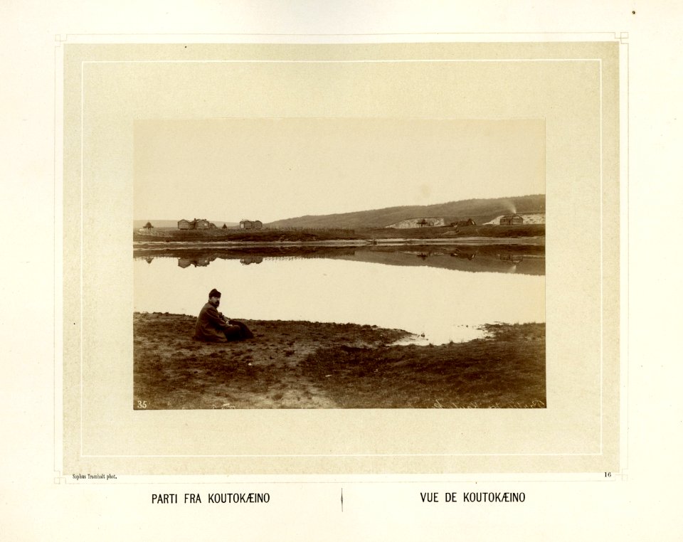 Mann sitter ved elvebredden i Kautokeino. Bebyggelse i bakgrunnnen - Norsk folkemuseum - NF.15006-017