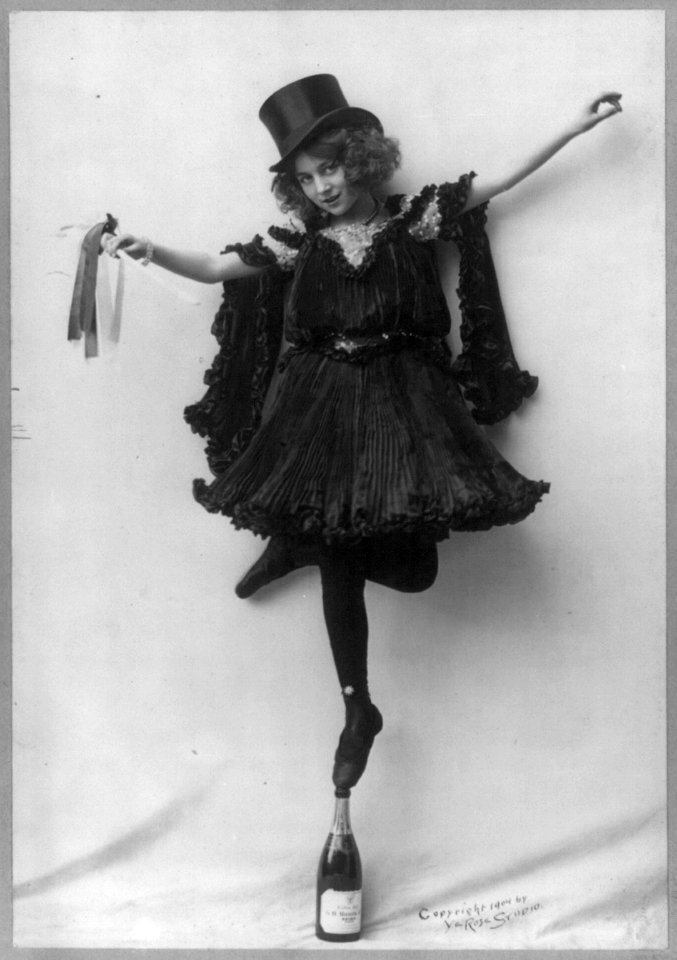 Model posed wearing black pressed pleats, top hat, and ballet shoes, standing tip-toe on champagne bottle LCCN2004678788