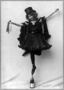 Model posed wearing black pressed pleats, top hat, and ballet shoes, standing tip-toe on champagne bottle LCCN2004678788