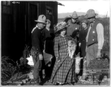 Mary Pickford, 1893-, full length portrait, standing, facing right, in front of building, holding dog, in movie Rags (western), with 3 men and 2 women - Rags Farewell to the West LCCN2005689188