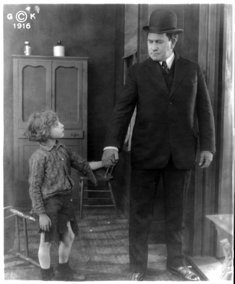 Motion picture still showing man wearing derby and holding hand of boy wearing cutoff trousers. LCCN2002699486