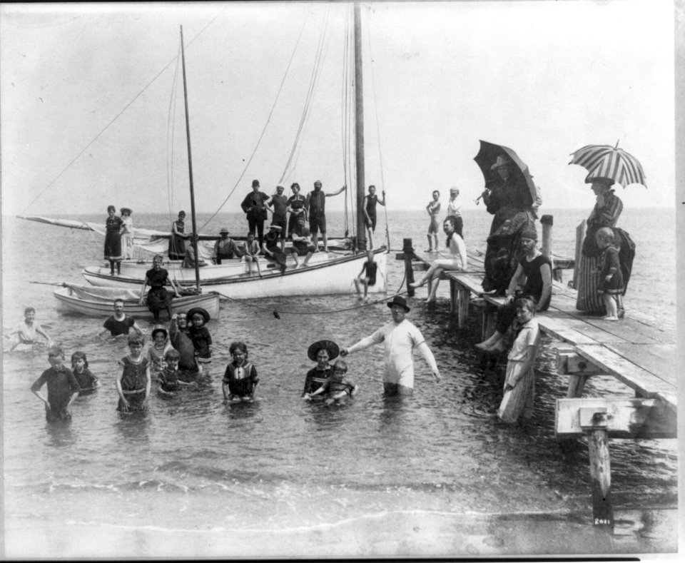 Men, women, and children in bathing suits on dock, sailboats, and in water; Frances Benjamin Johnston sits on edge of small catboat LCCN2005688906