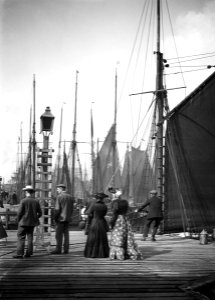 Men and women onlookers at the end of the quay looking at the forest of masts and sails in the harbour. RMG P27510